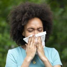 An African-American Woman with Sinusitis Problems is Feeling Displeased and Blowing Nose in Napkin During a Walk in City Park During a Summer Day.